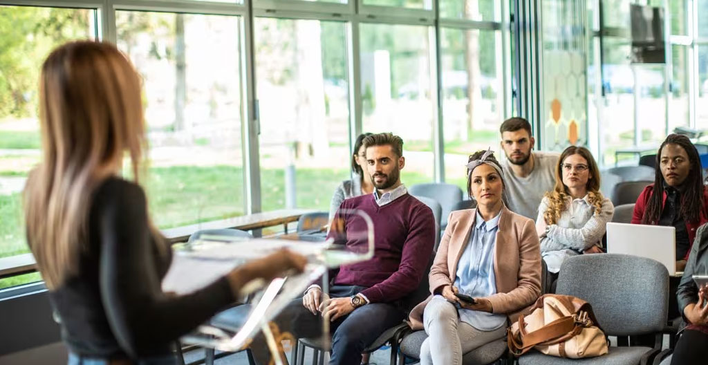 Orateur debout face à une salle, posture ouverte, dos droit, regard dirigé vers l'auditoire avec une expression calme et assurée